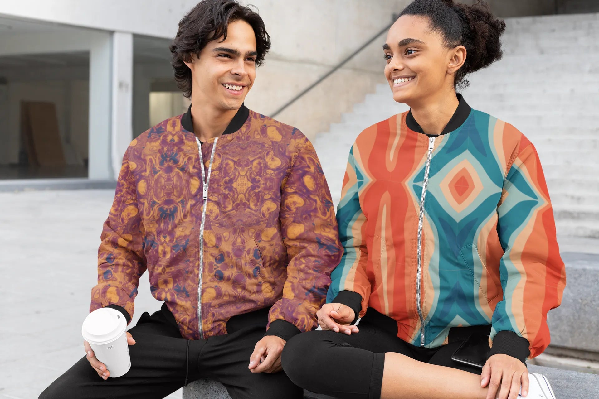 Two people wearing colorful patterned bomber jackets sitting outdoors.